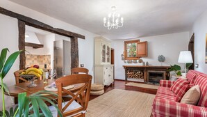 Un'area pranzo con un tavolo di legno, sedie e una ciotola di frutta. Si nota un mobiletto bianco, un divano rosso e un lampadario.