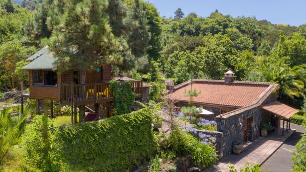 Una baita di legno con un balcone, immersa nel verde e affiancata da un edificio in pietra con tetto in tegole.