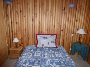 Quarto com parede de ripas de madeira, cama com colcha azul e branca estampada, duas luminárias de cabeceira e uma mesa pequena com toalha de mesa verde.