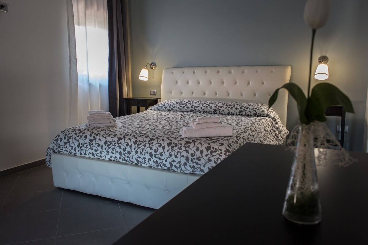 A bedroom with a tufted headboard, a patterned bedspread, and neatly folded towels on the bed. There is a bedside table with a lamp and a vase with a plant.