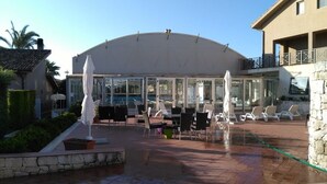 A covered outdoor seating area with tables and chairs, a fountain, and a glass-enclosed building in the background. - Hotel La Cavalera (Augusta)