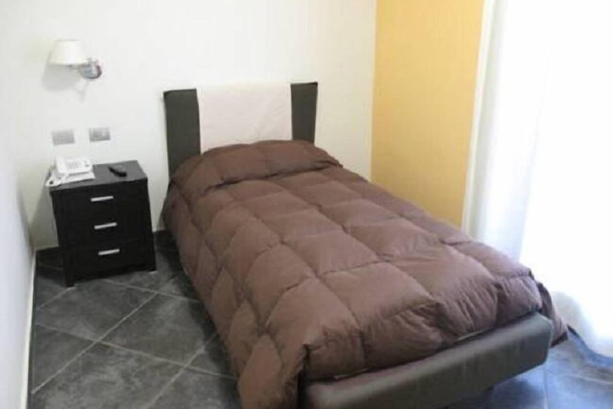 A single bed with a brown quilt, a nightstand with drawers, a lamp, and a plain wall.
