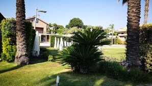 A well-maintained garden with palm trees, a lawn, and a building in the background. - Hotel La Cavalera (Augusta)