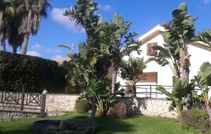 A white building with a stone wall and a metal fence. There are large green plants and a small tree in front of the building. - Hotel La Cavalera (Augusta)
