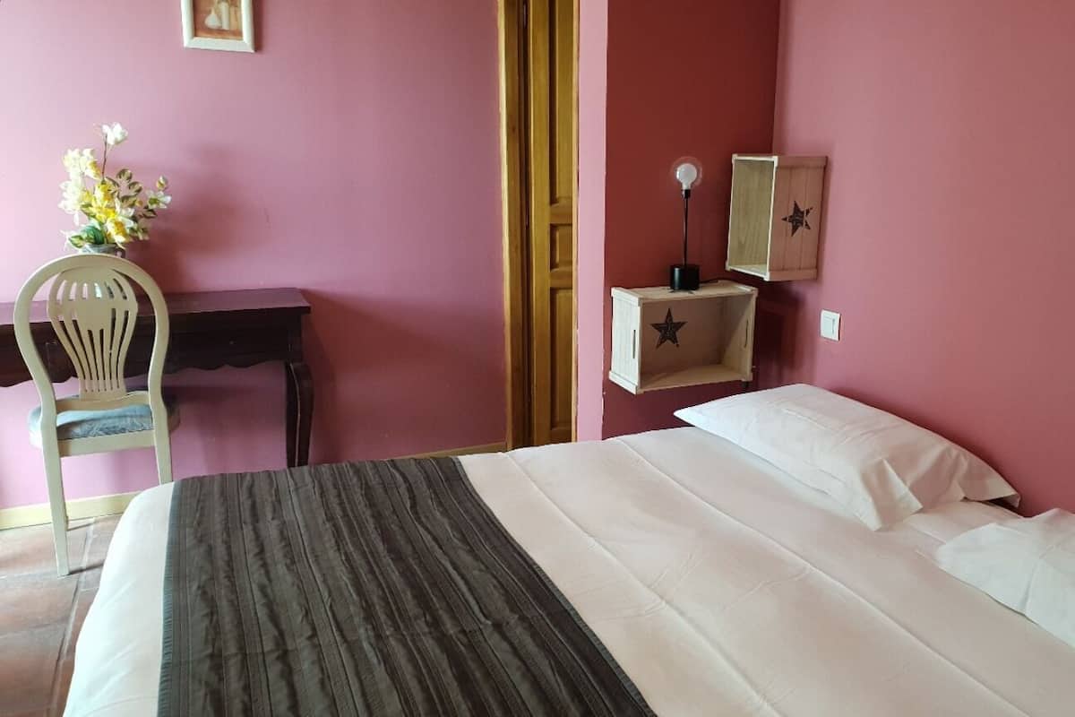 A bedroom with a bed, a desk, a chair, a nightstand, and a wall-mounted lamp.