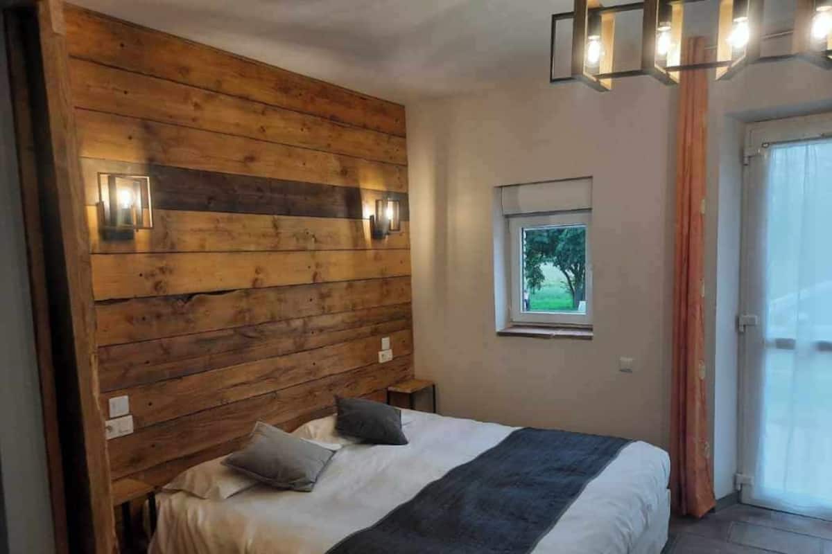 A bedroom with a wooden headboard, a bed with pillows, a nightstand, a window with a view of greenery, and a door leading to a balcony.