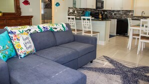 A modern living room with a gray sectional sofa, white chairs, and a kitchen in the background.