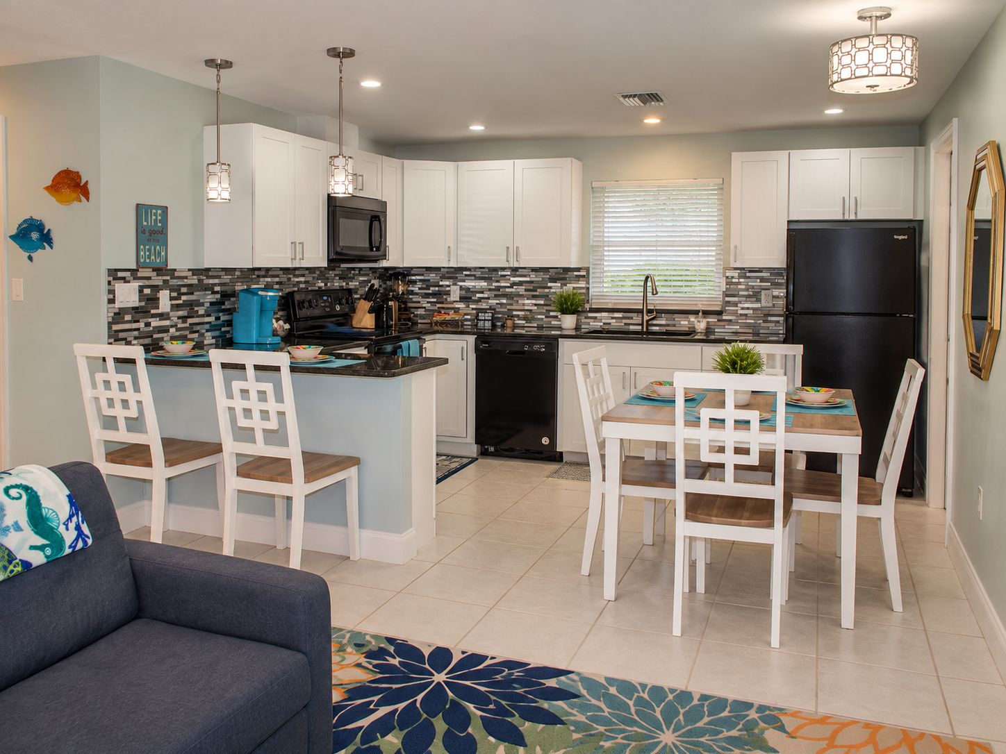 A modern kitchen with white cabinets, a black refrigerator, and a dining area with white chairs and a blue rug.