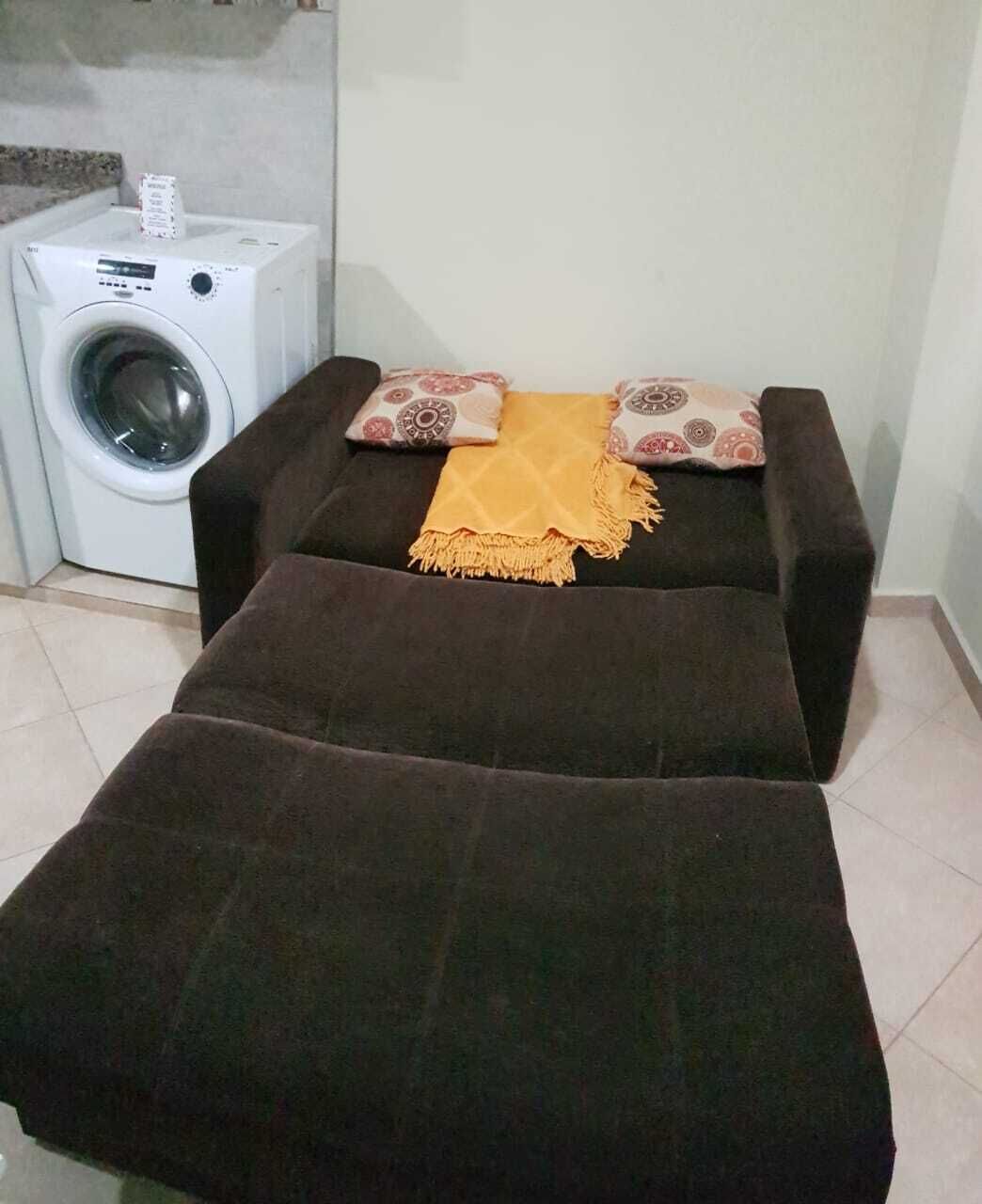 Een kleine, donkere bank met een gele deken en gekleurde kussens in een kamer met een wasmachine.
