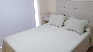 En pænt redt seng med hvidt sengegavl og puder, et natbord med lampe, og en hvid dør.
