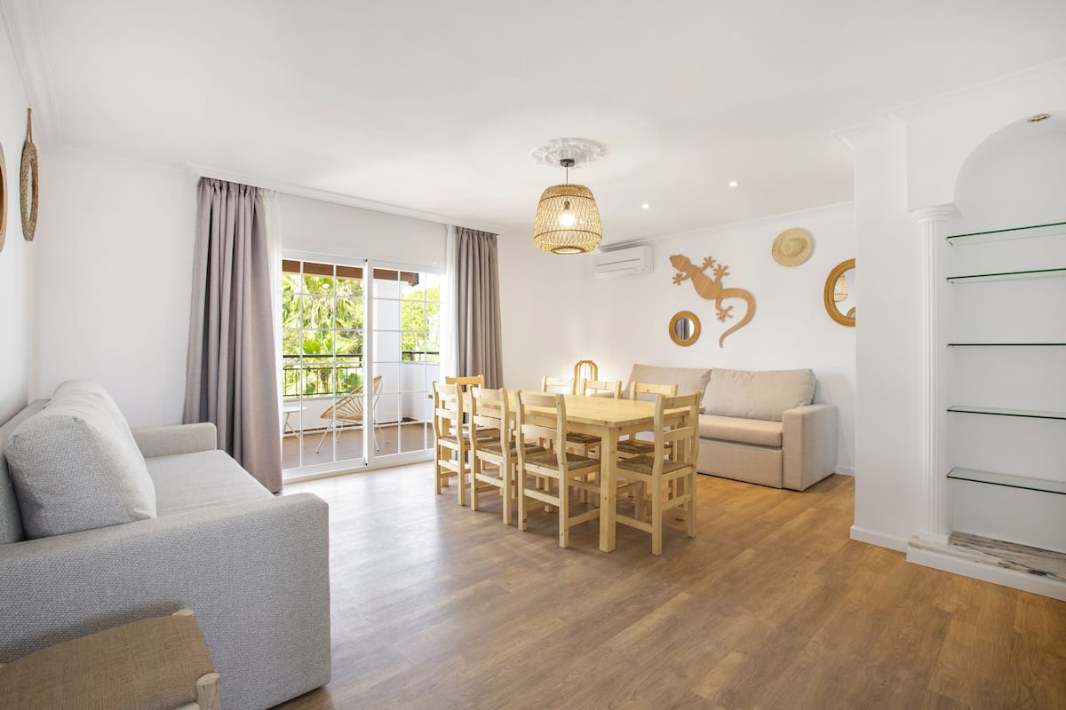 A spacious living area with a sofa, dining table, and chairs, a balcony with a view, and decorative wall art.