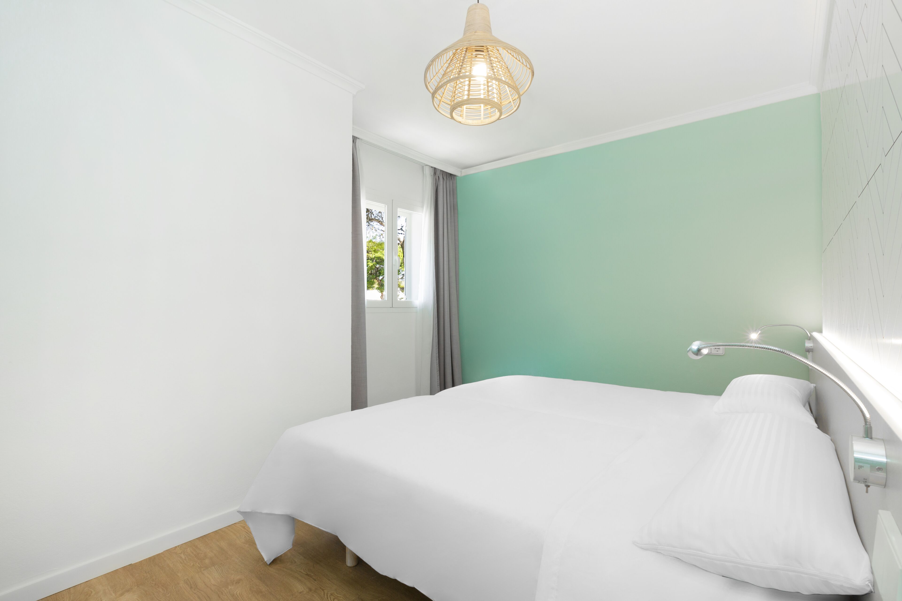 A bedroom with a bed, a bedside table, a lamp, a window with curtains, and a wall-mounted light fixture.