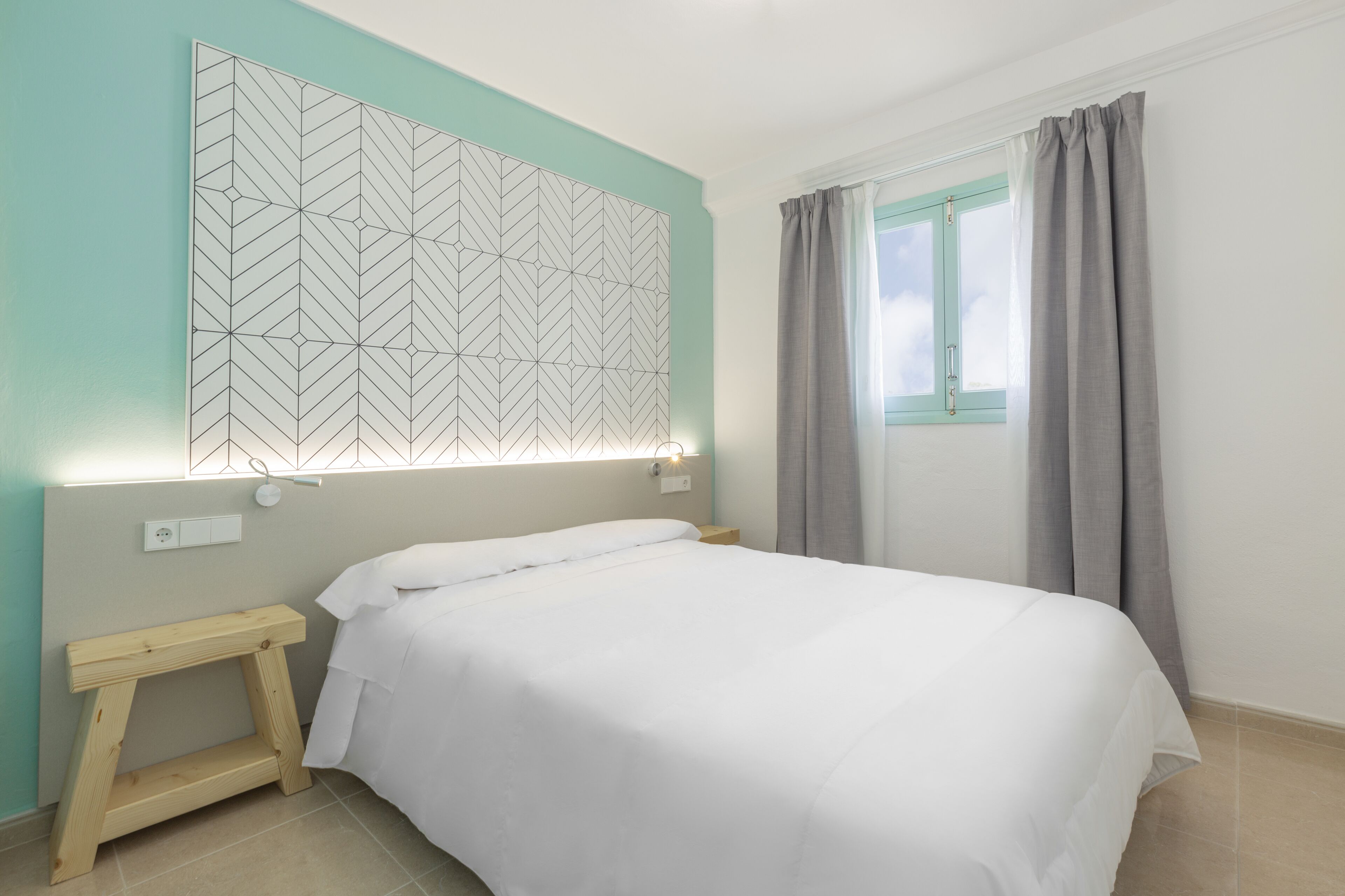 Un dormitorio ordenado con cama, mesita de noche, luz en la pared y ventana con cortinas.