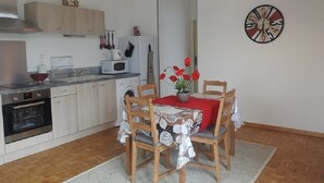 A compact kitchen with wooden cabinets, a microwave, and a sink. There is a dining table with chairs and a vase of red flowers. A clock is on the wall.