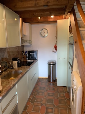 A compact kitchen with white cabinets, a stainless steel sink, a Nespresso machine, a trash can, and a washing machine.