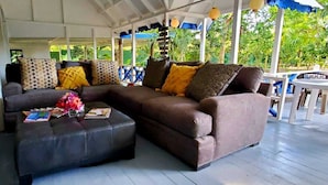 A covered outdoor seating area with a brown sofa and ottoman, surrounded by blue and white chairs. - Bak A Yaad Overt Green Cottage (Negril)