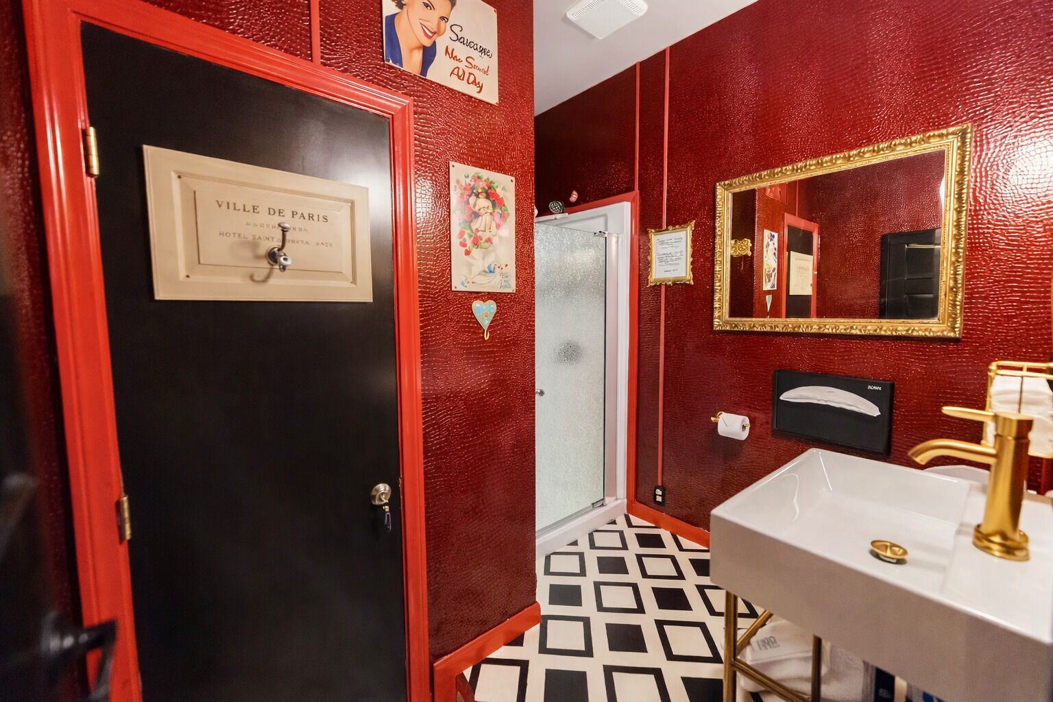 Um banheiro com parede vermelha, porta preta, pia branca com detalhes dourados e um espelho com moldura dourada.
