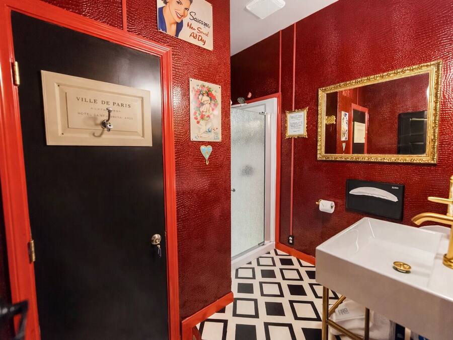 Um banheiro com parede vermelha, porta preta, pia branca com detalhes dourados e um espelho com moldura dourada.