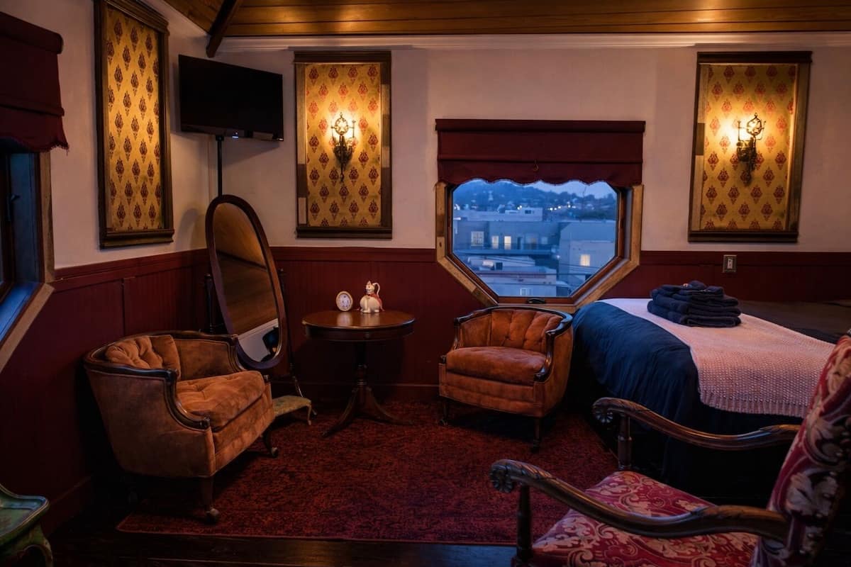 Une pièce avec une grande fenêtre, deux fauteuils, une petite table avec une horloge et un lit recouvert d’une couverture bleue.
