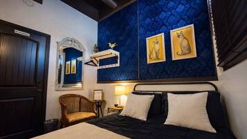 Una habitación de hotel con una puerta de madera oscura, una pared azul estampada, una cama con ropa de cama blanco y negro, una silla de mimbre y dos cuadros con imágenes de gatos.