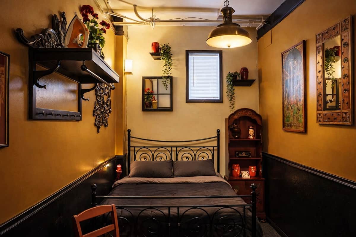 Une petite pièce comprenant un lit, une chaise, une étagère avec des objets décoratifs, un luminaire suspendu et une fenêtre avec des stores.