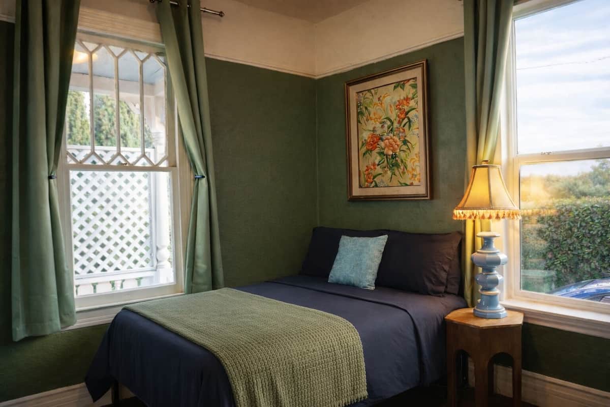 Une chambre à coucher avec un lit, une table de chevet, une lampe, une fenêtre avec des rideaux et un tableau floral accroché au mur.