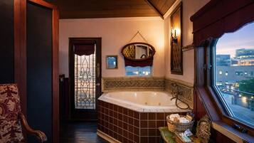 Un baño con una bañera grande, una silla de madera, un ventanal con vista a la ciudad y un espejo decorativo.