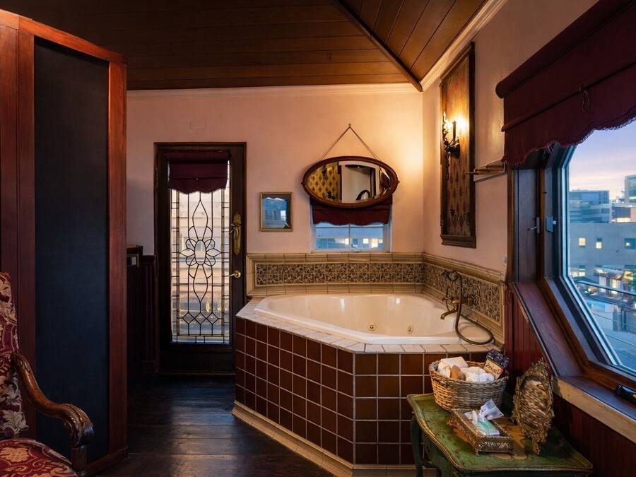 Un baño con una bañera grande, una silla de madera, un ventanal con vista a la ciudad y un espejo decorativo.