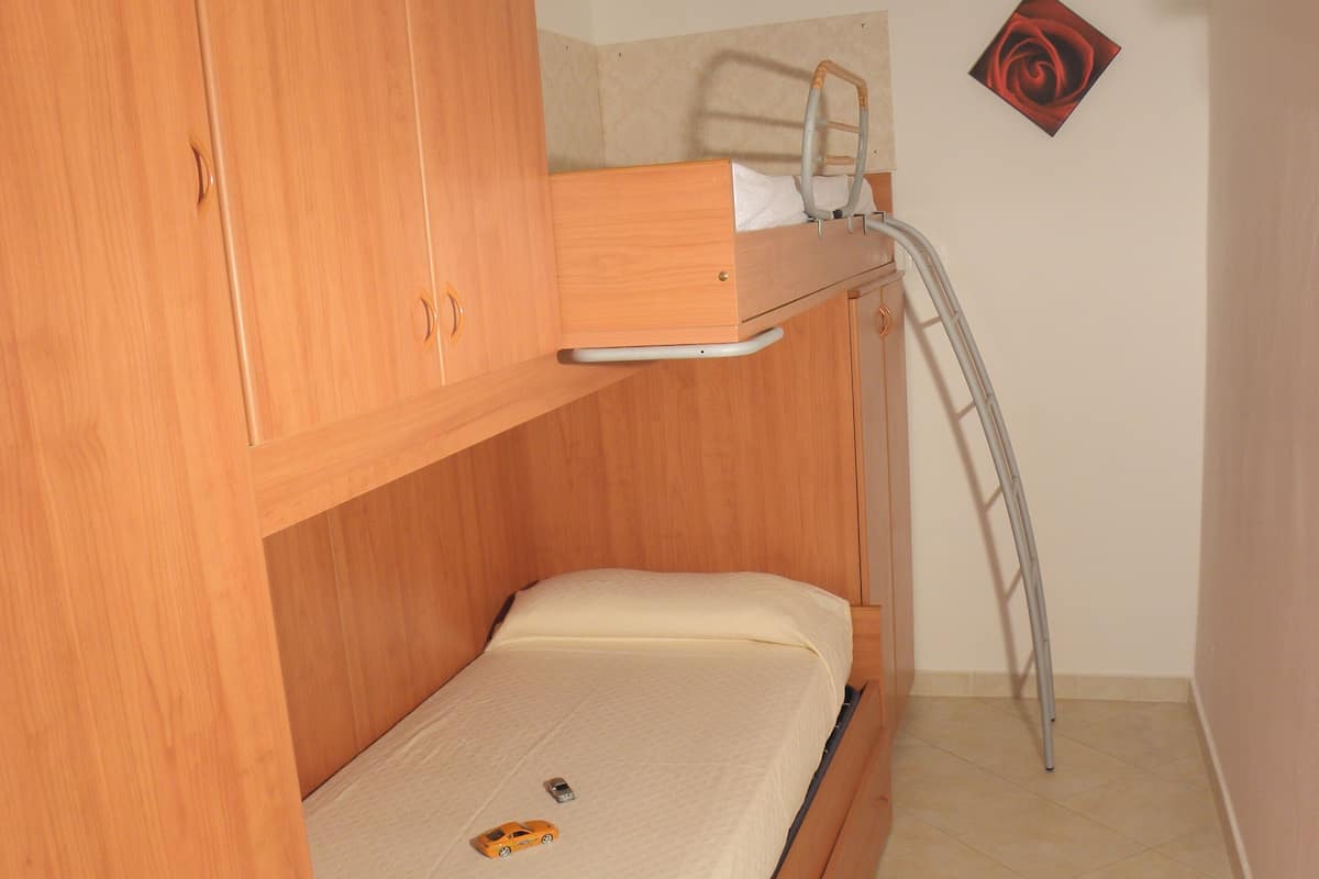 A small, compact bedroom with a bunk bed, a wooden wardrobe, a small bedside table, and a single bed.