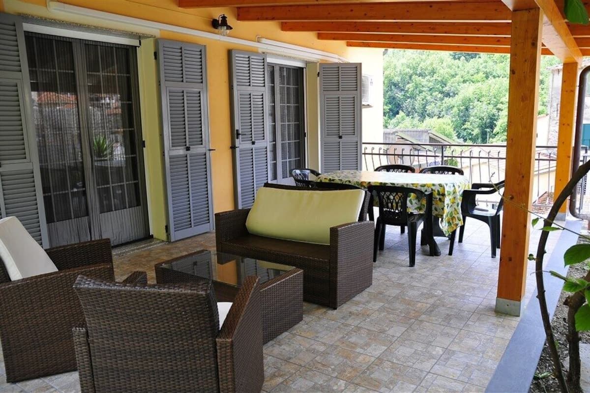 A covered outdoor seating area with wicker furniture, a table with a floral tablecloth, and a view of greenery in the background.