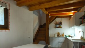 Une cuisine avec des poutres en bois, un escalier, un coin repas avec une table et des chaises, et une étagère contenant des ustensiles de cuisine.