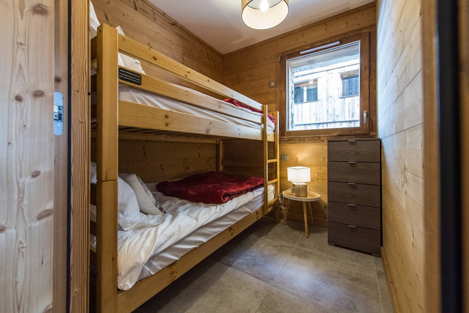 Puinen mökkihuone, jossa on kerrossängyt, pieni pöytä lampun kanssa ja ikkuna, josta on näkymä rakennuksiin.