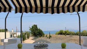 A table with a bowl and apples under a striped awning, overlooking a beach and dry landscape. - Καλώς ήρθατε στην εξοχική κατοικία στην Αίγινα (Aegina)