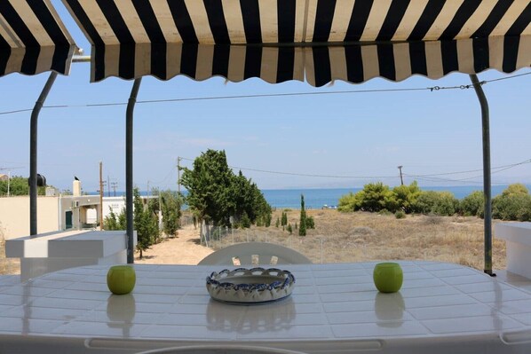 A table with a bowl and apples under a striped awning, overlooking a beach and dry landscape.