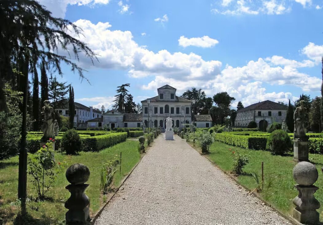 Ein gepflegter Gartenweg führt zu einem klassischen Gebäude mit symmetrischer Bepflanzung und Statuen.