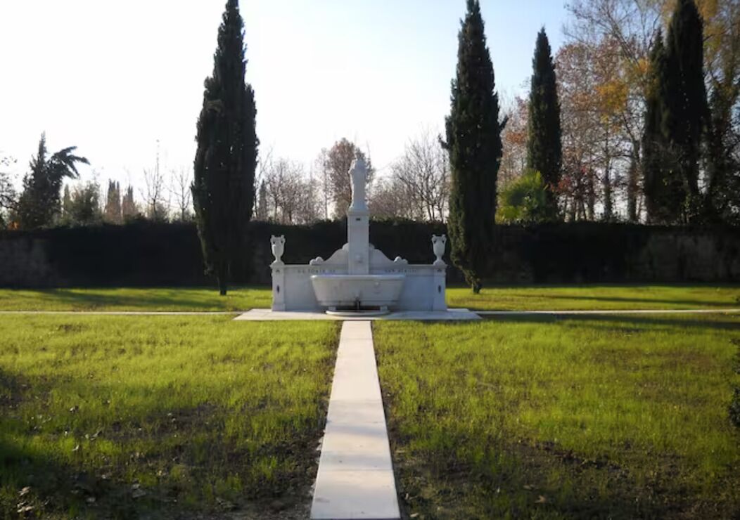 A symmetrical garden with a central fountain, surrounded by neatly trimmed grass and tall trees.