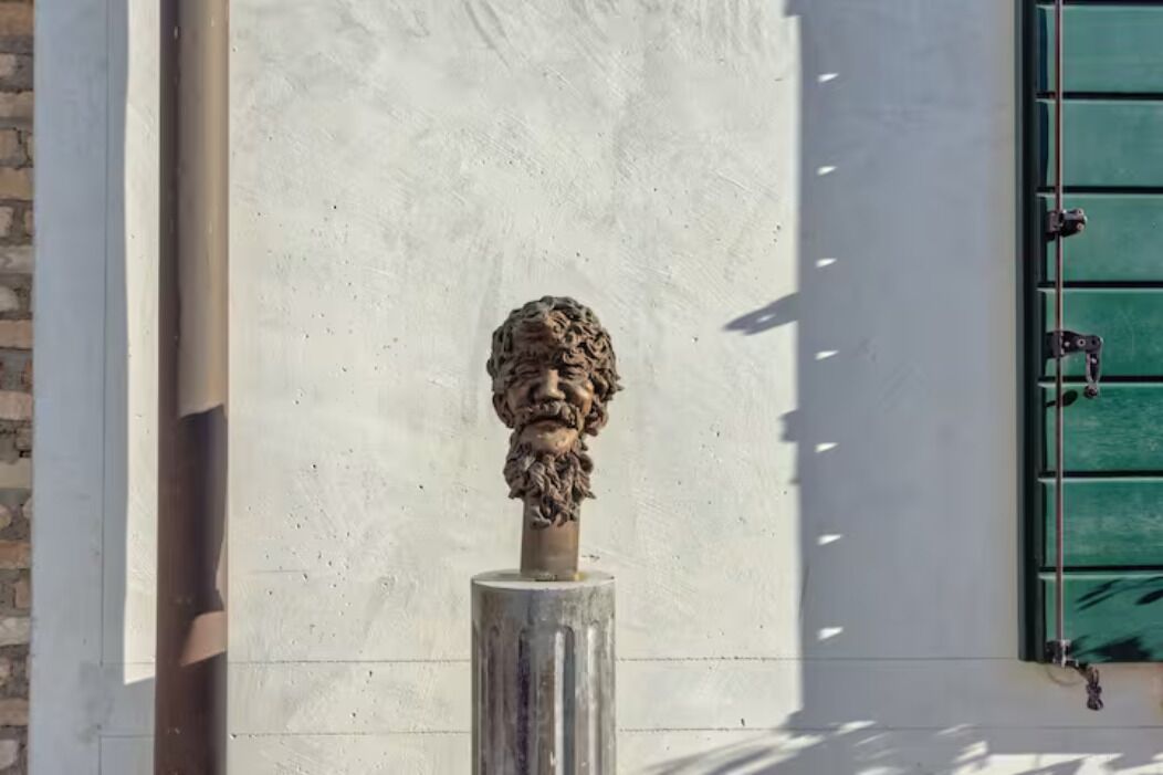 Un buste en pierre, posé sur un piédestal, se dresse devant un mur blanc orné d’un volet vert.