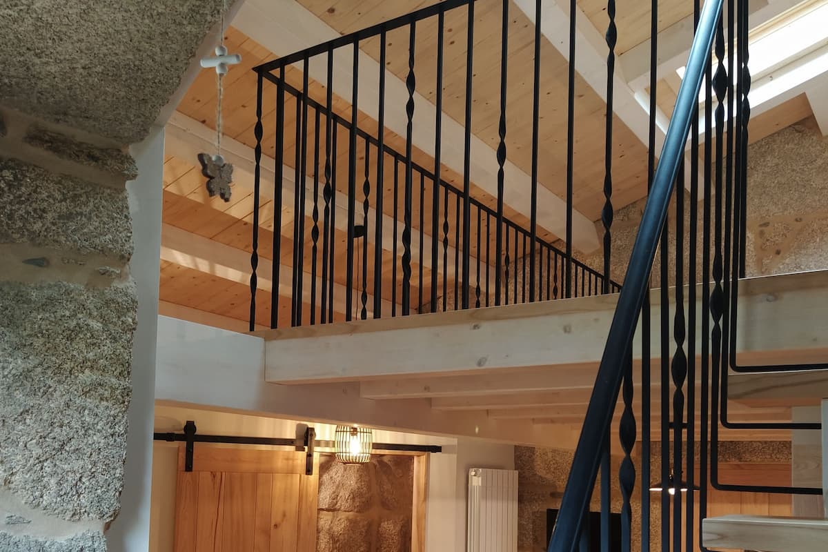 A loft bedroom with a wooden ceiling, a stone wall, a bed with patterned bedding, a hanging light fixture, and a spiral staircase.