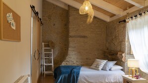 Une chambre avec un mur en pierre, un lit recouvert d’une couverture bleue, une échelle en bois et un luminaire suspendu.