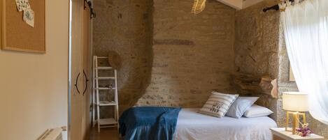 A bedroom with a stone wall, a bed with a blue blanket, a wooden ladder, and a hanging light fixture.