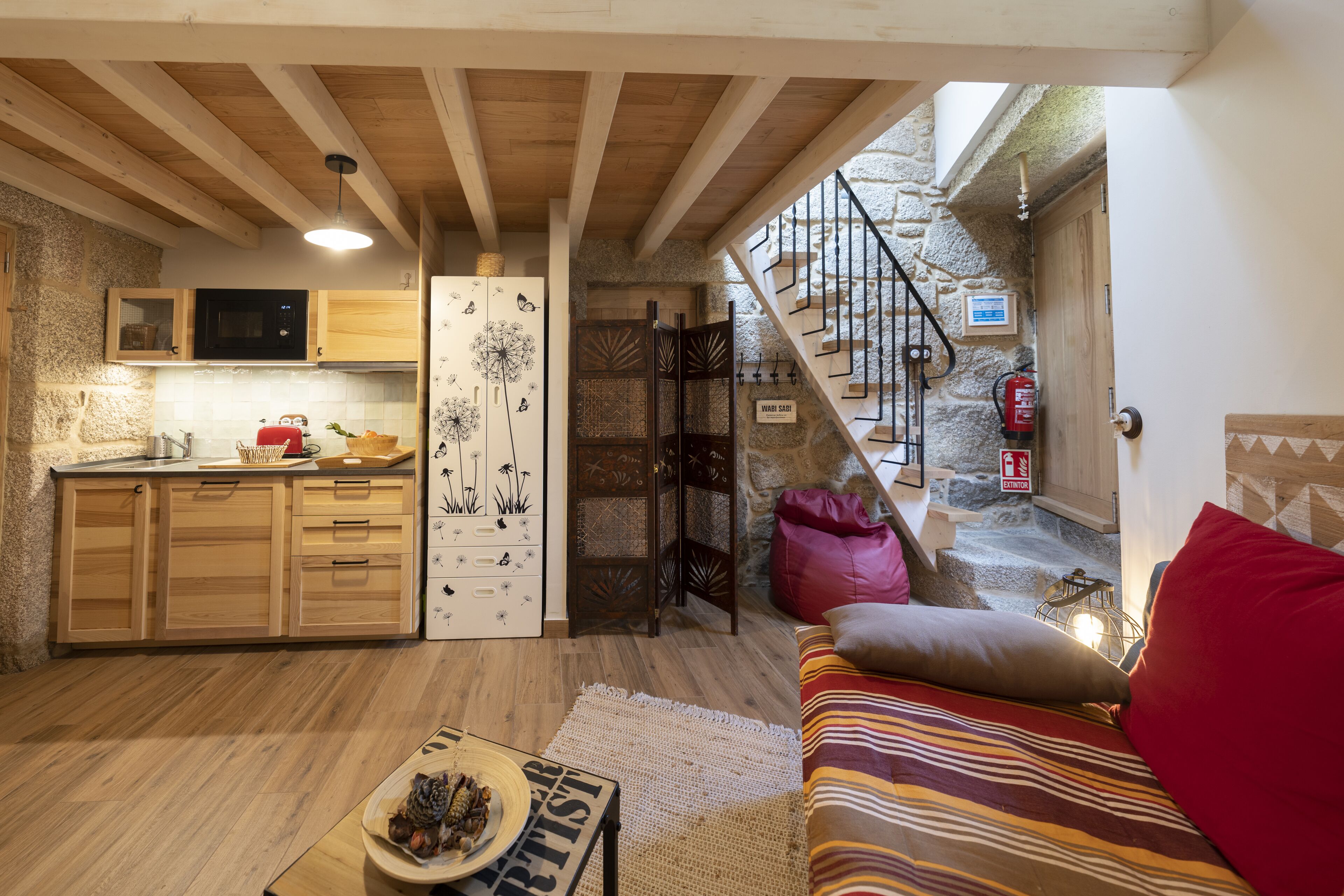 A cozy kitchen with wooden cabinets, a microwave, and a dandelion-themed door. A staircase leads to a stone-walled area with a fire extinguisher.