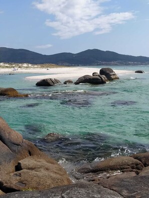 Una playa con agua turquesa cristalina, rocas grandes y montañas a lo lejos.