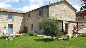 Un bâtiment en pierre avec des volets bleus, un jardin où poussent différentes espèces de plantes et des sièges installés à l’extérieur.