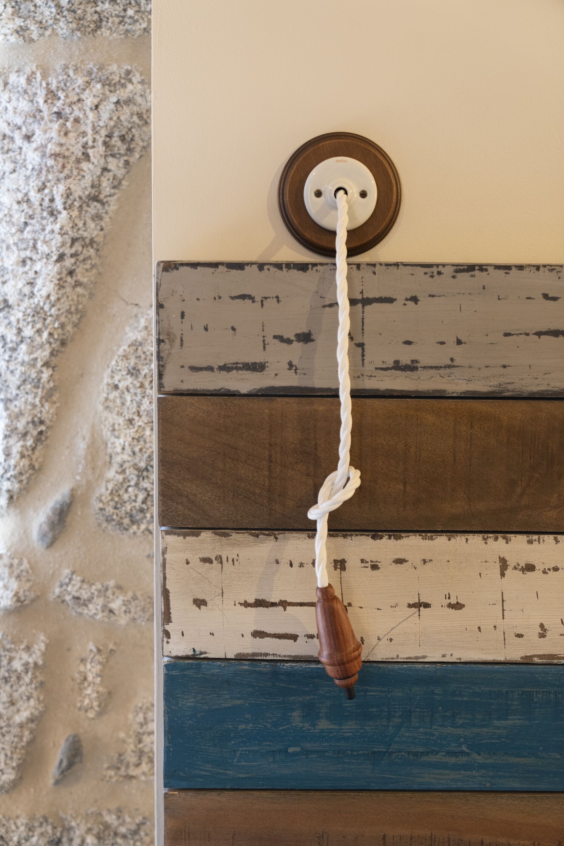 A rustic wall with horizontal wooden planks in varying shades of brown and blue, a white rope hanging from a wooden hook, and a textured stone wall to the left.