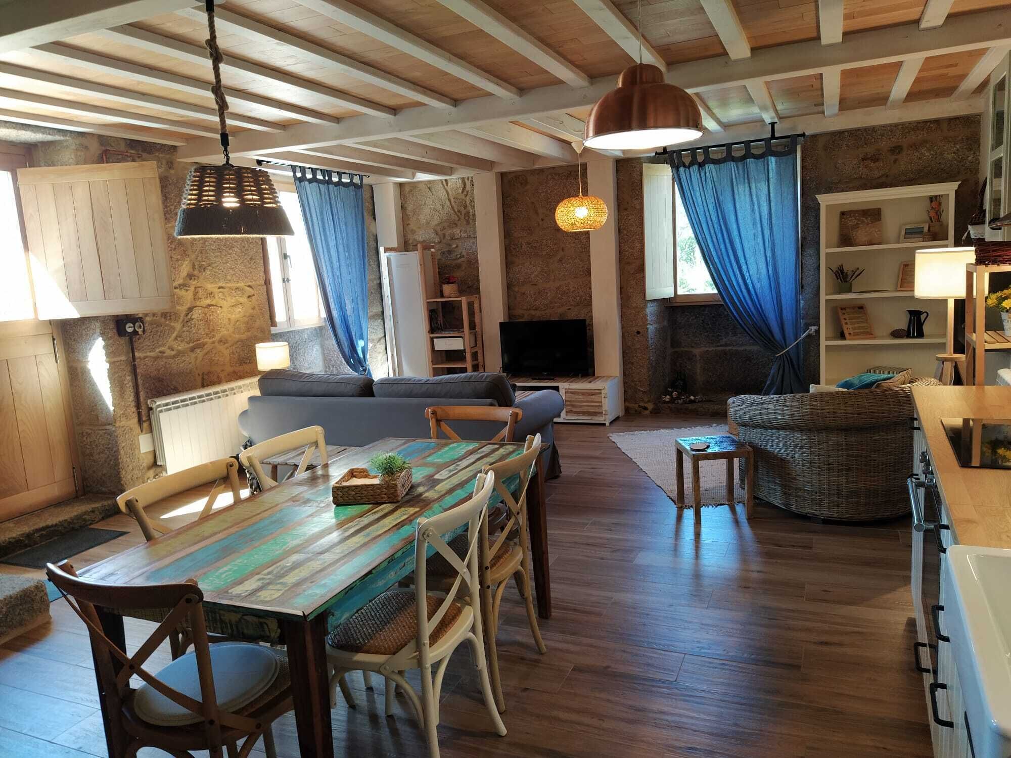 A cozy living room with a wooden ceiling, a dining table with chairs, a sofa, and a television.