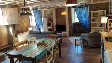 A cozy living room with a wooden ceiling, a dining table with chairs, a sofa, and a television.