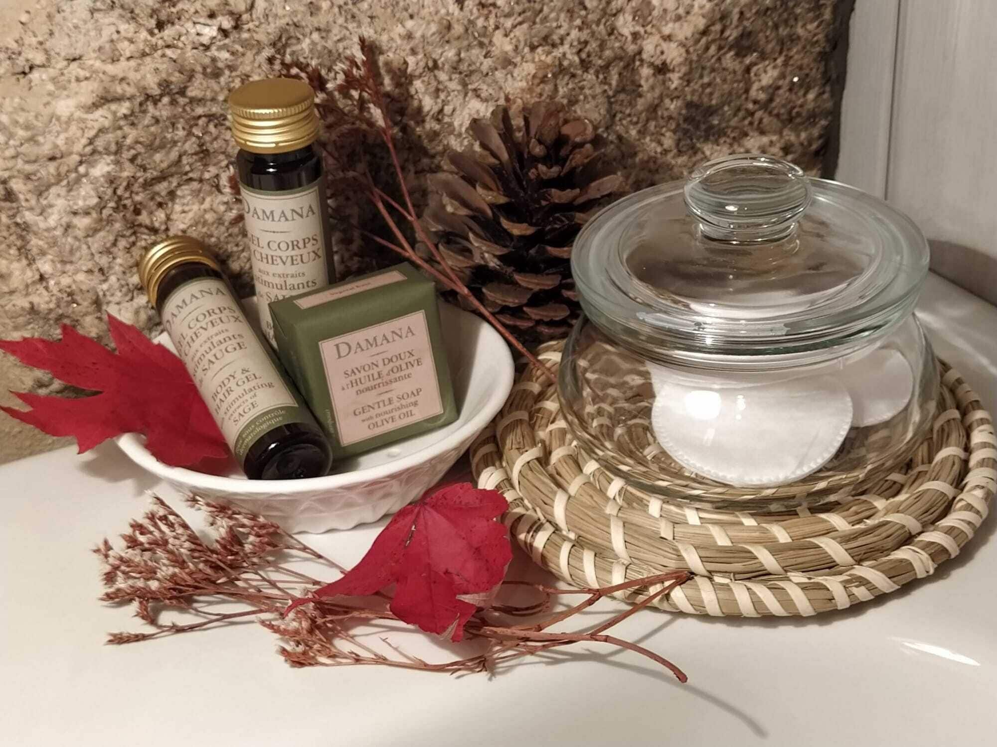 A collection of Damana body care products including a body lotion, a body wash, and a soap, along with a glass jar containing a white substance, possibly a lotion or cream, set on a woven mat.