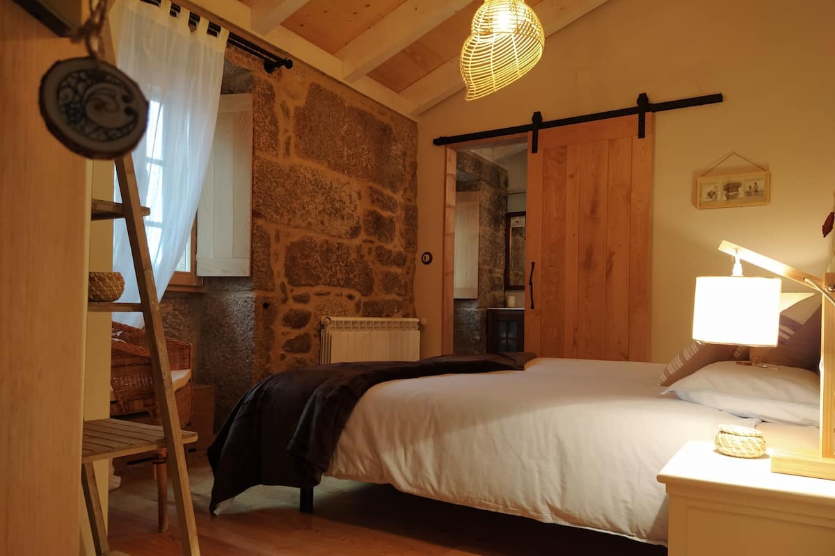 A bedroom with a bed, a bedside lamp, a wooden ladder, a stone wall, and a wooden ceiling.