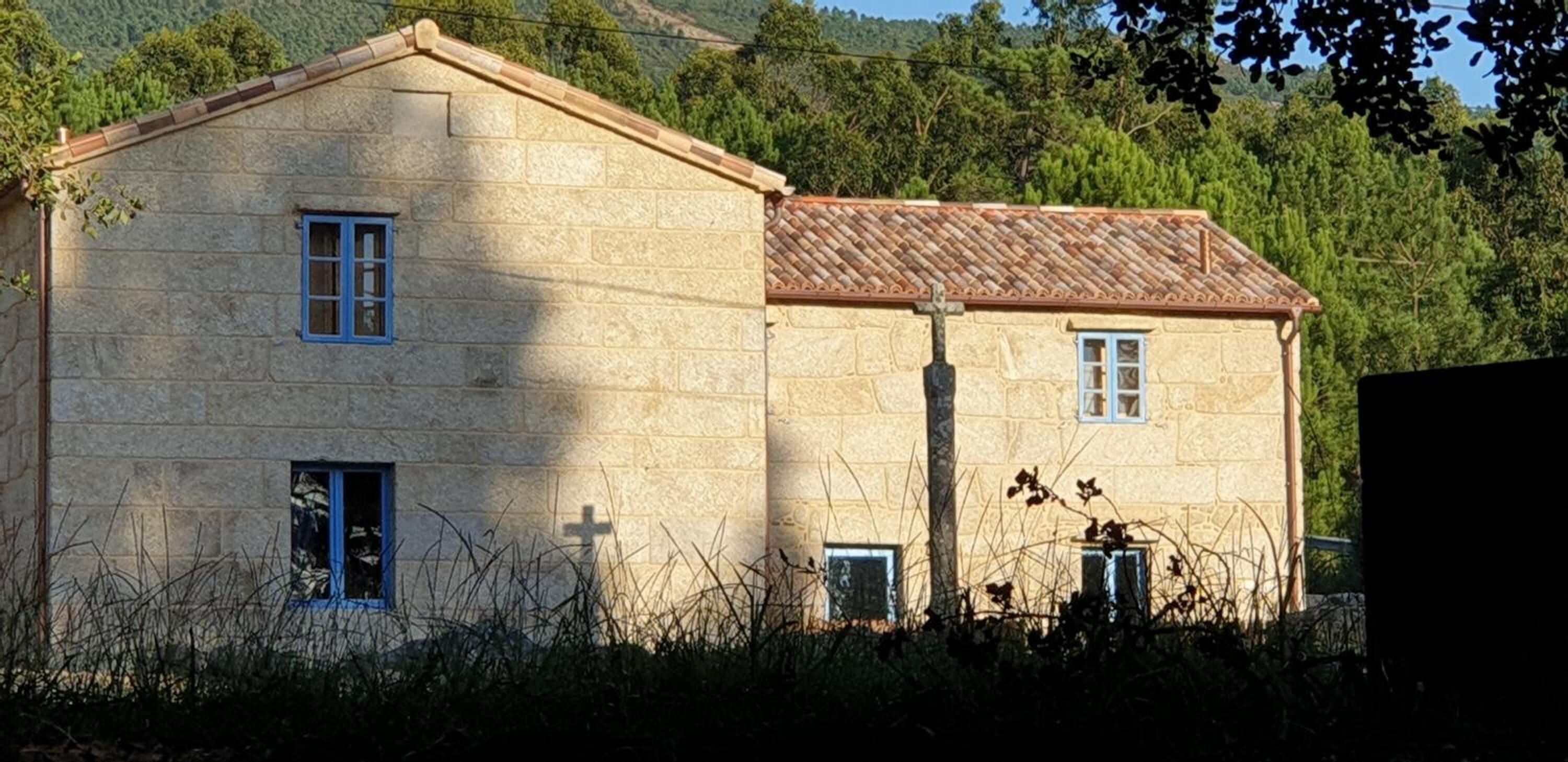 A stone building with a tiled roof, two windows, and a cross on the wall.