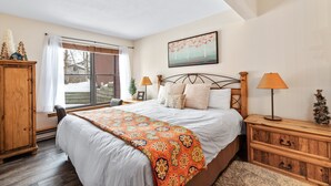 A bedroom with a large bed, wooden headboard, two bedside lamps, a nightstand, a small Christmas tree, and a window with a view of trees. - Family-Friendly Comfort at Timber Run – 2-Bedroom Condo Near the Slopes! (Steamboat Springs)
